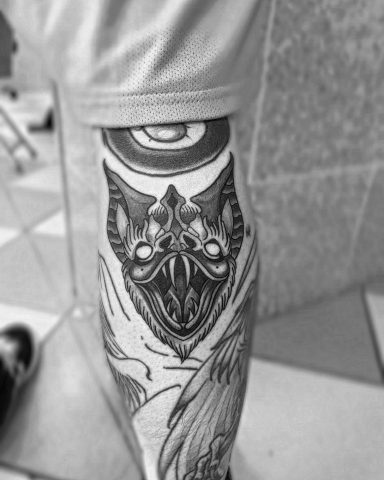 Tattoo auf einem Bein mit einem detaillierten, maskenartigen Design in Schwarzweiß.