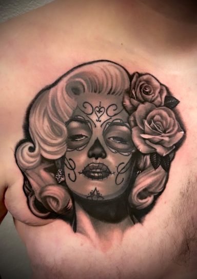 Tattoo einer Frau mit Blumen im Haar und Totenkopf-Makeup auf der Brust.