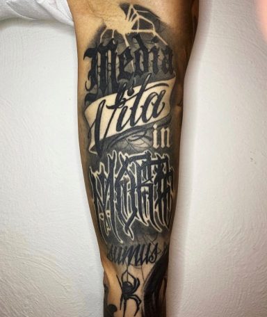 Tattoo auf dem Unterarm mit den Worten "Media Vita in Morte" und verschiedenen Grafikelementen.
