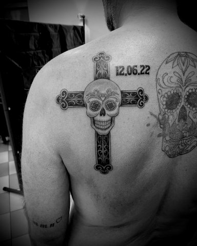 Kreuztattoo mit Totenkopf und Datum 12.06.22 auf dem Rücken einer Person.