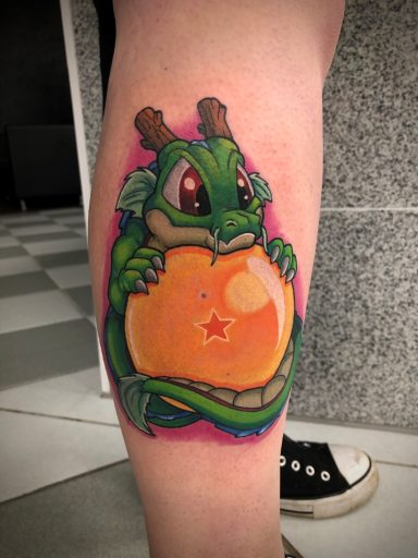 Ein grüner Drache hält eine orangefarbene Kugel mit einem Stern, auf einem Bein tätowiert.